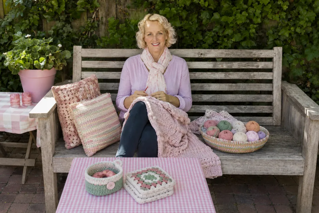 About 1 Amanda Parker sitting on a wooden garden bench crocheting a soft pink blanket surrounded by pastel yarn, crochet pillows, and small handmade crochet projects outdoors.